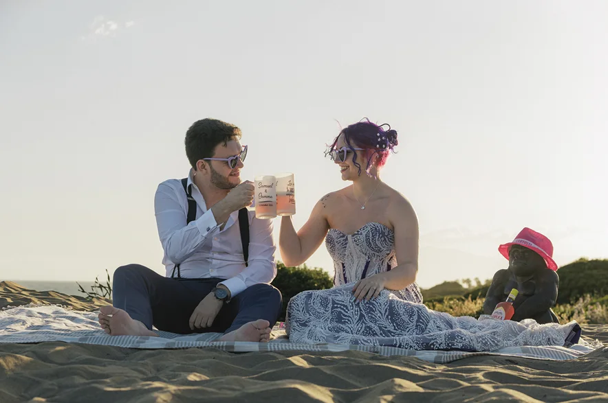 Una pareja brinda sentada en una manta sobre la arena, con el mar detrás.