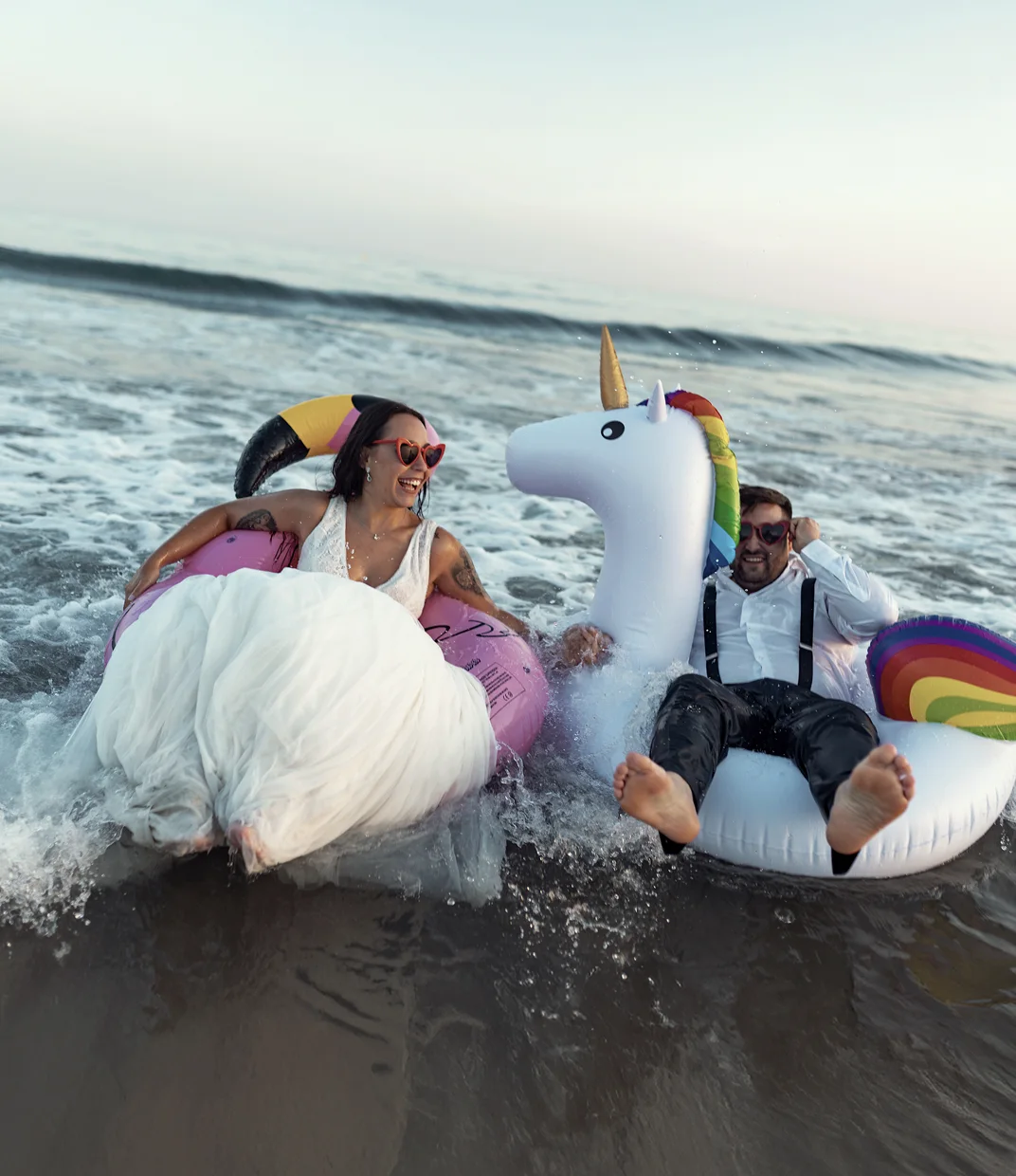 Una pareja recién casada, con el vestido empapado, se ríe dentro del mar sobre un flotador de tucán y otro de unicornio.