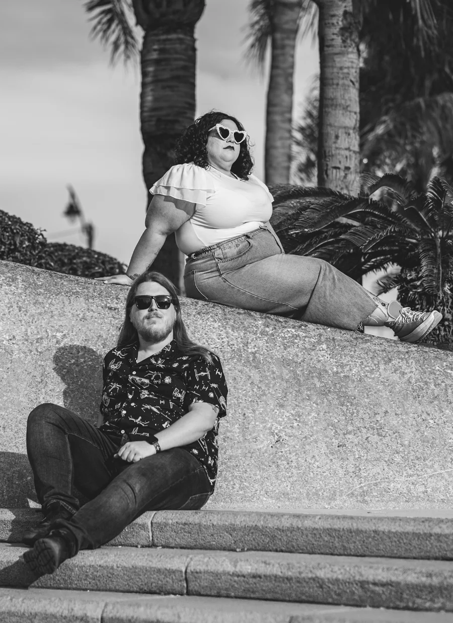 Tom y Paula, con gafas de sol, sentados en un muro de piedra bajo unas palmeras. En blanco y negro.