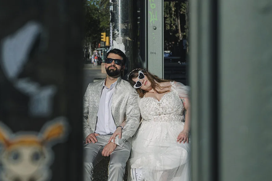 Él con traje de lentejuelas y ella de novia, los dos con gafas de sol, recostados en un portal.